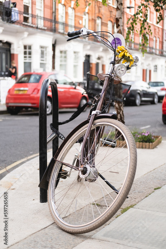 Vertical photo of a bicycle with a yellow sunflower flower on the handlebars. The bike has a bell, a basket and light and is parked on the street. Urban sustainable transport. Red car the background.