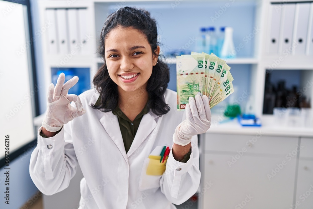Hispanic woman with dark hair working at scientist laboratory holding money doing ok sign with fingers, smiling friendly gesturing excellent symbol Hispanic woman with dark hair working at scientist laboratory holding money doing ok sign with fingers, smiling friendly gesturing excellent symbol