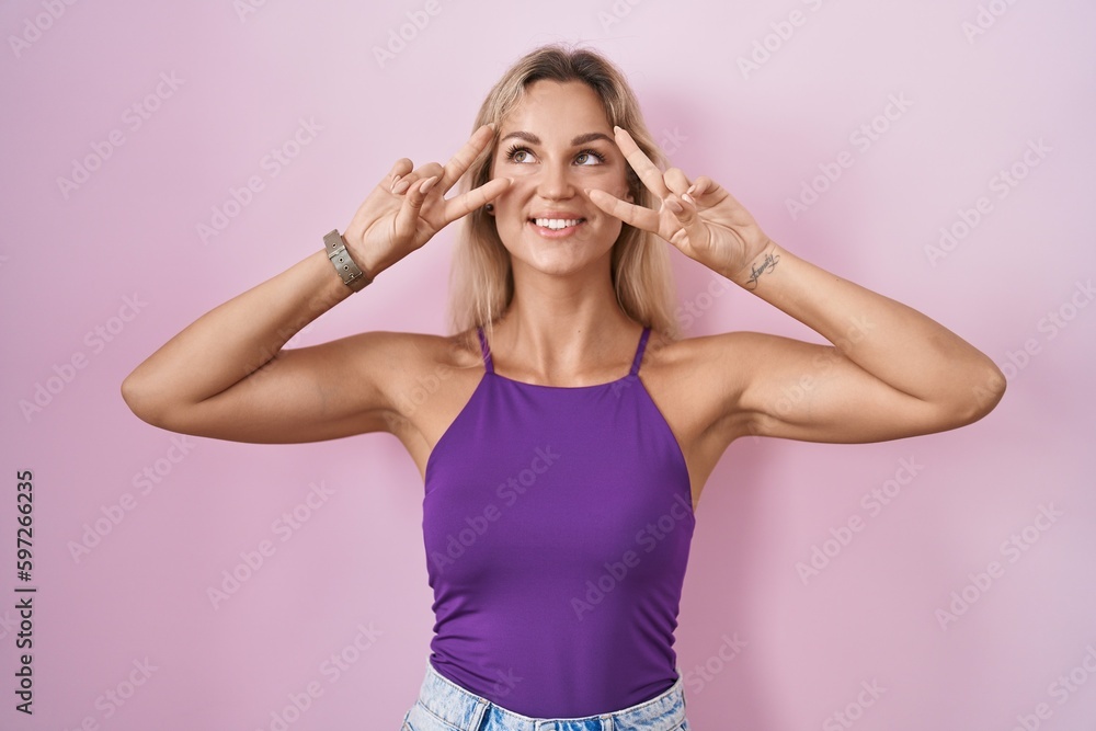 Young blonde woman standing over pink background doing peace symbol with fingers over face, smiling cheerful showing victory Young blonde woman standing over pink background doing peace symbol with fingers over face, smiling cheerful showing victory