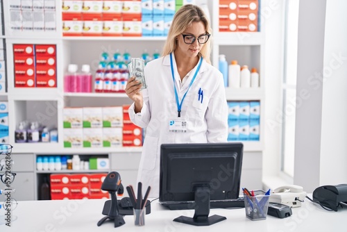 Young blonde woman pharmacist holding dollars using computer at pharmacy