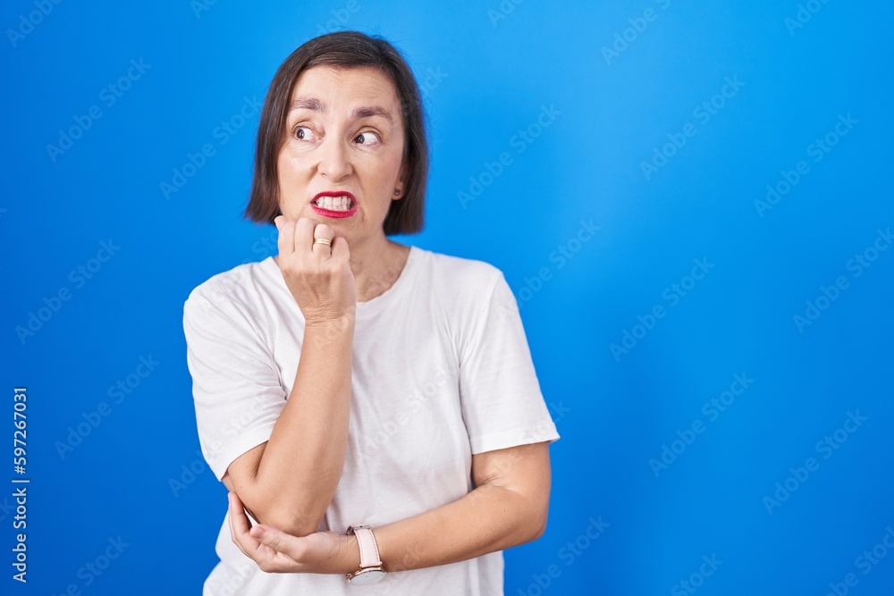 Middle age hispanic woman standing over blue background looking stressed and nervous with hands on mouth biting nails. anxiety problem. Middle age hispanic woman standing over blue background looking stressed and nervous with hands on mouth biting nails. anxiety problem.