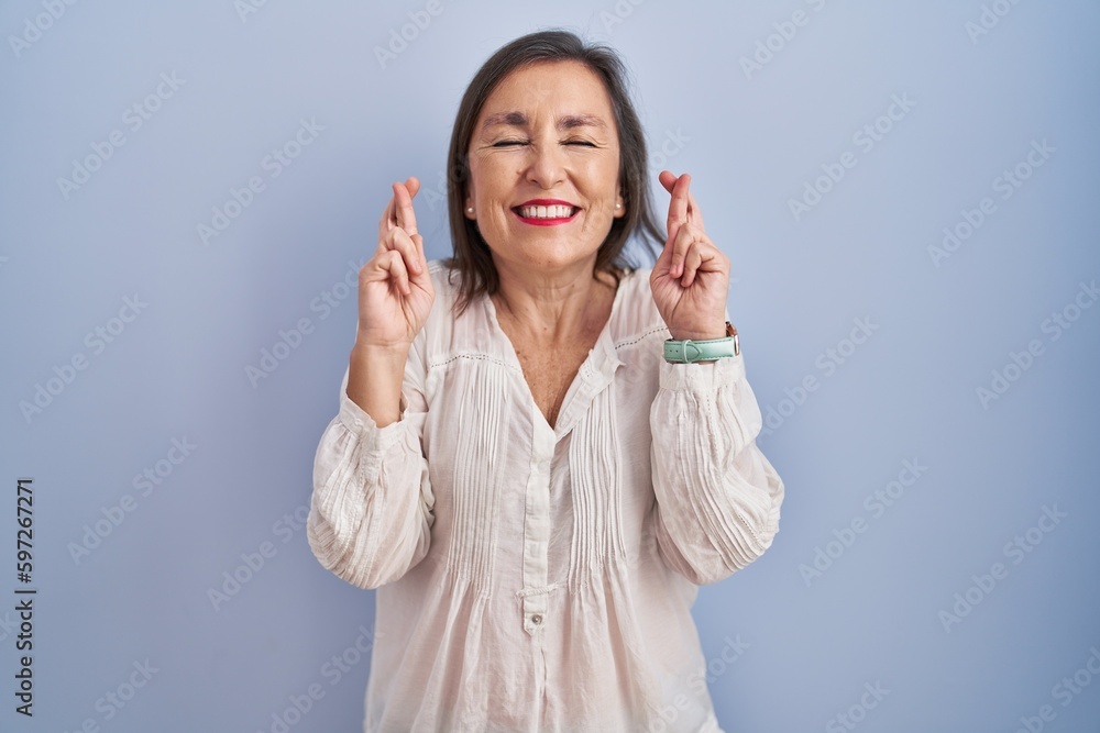 Middle age hispanic woman standing over blue background gesturing finger crossed smiling with hope and eyes closed. luck and superstitious concept.
