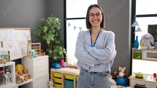 Obraz Young beautiful hispanic woman teacher smiling confident standing with arms crossed gesture at kindergarten