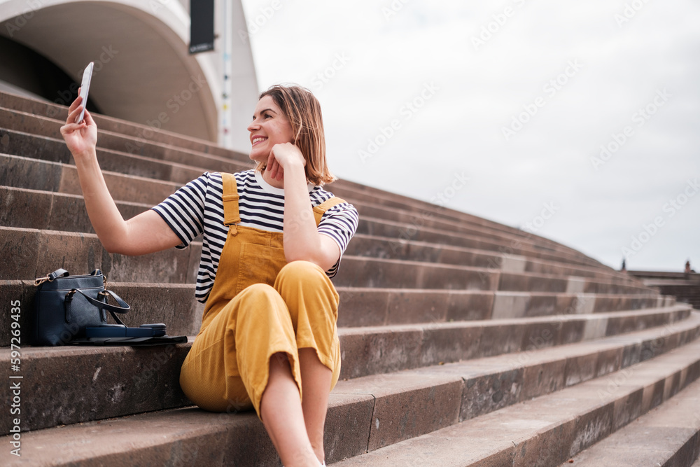 Young woman taking a selfie with her smartphone on some stairs outdoors. Concept: lifestyle, outdoors, smartphone