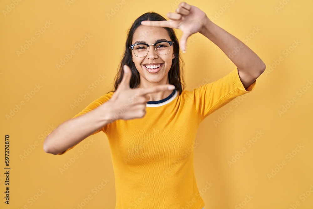 Fototapeta premium Young brazilian woman wearing glasses over yellow background smiling making frame with hands and fingers with happy face. creativity and photography concept.