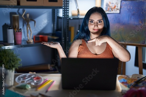 Young modern girl with blue hair sitting at art studio with laptop at night amazed and smiling to the camera while presenting with hand and pointing with finger.