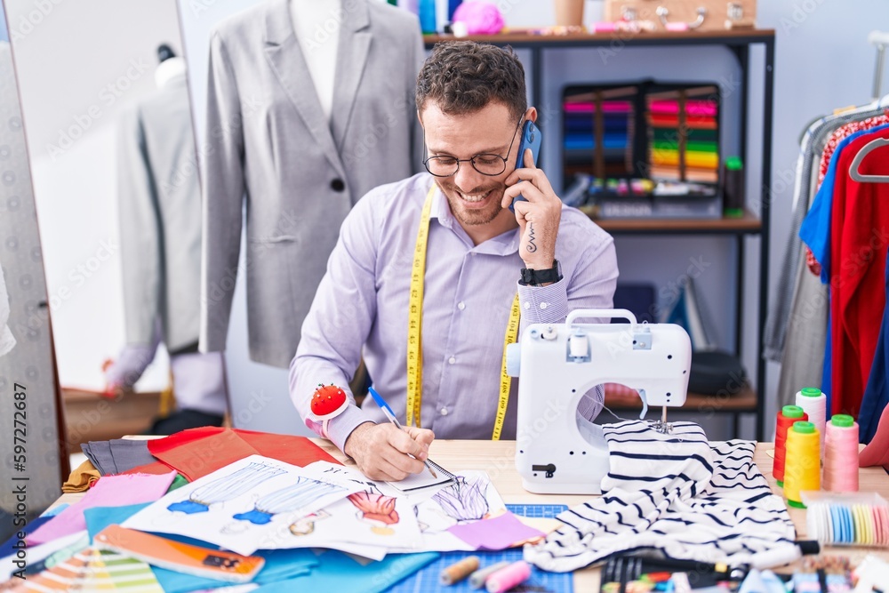 Young hispanic man tailor talking on smartphone writing on notebook at tailor shop