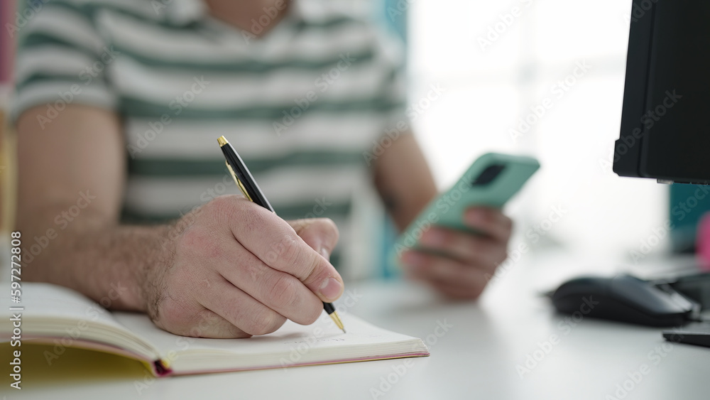 Young caucasian man using computer taking notes with smartphone at library university Young caucasian man using computer taking notes with smartphone at library university