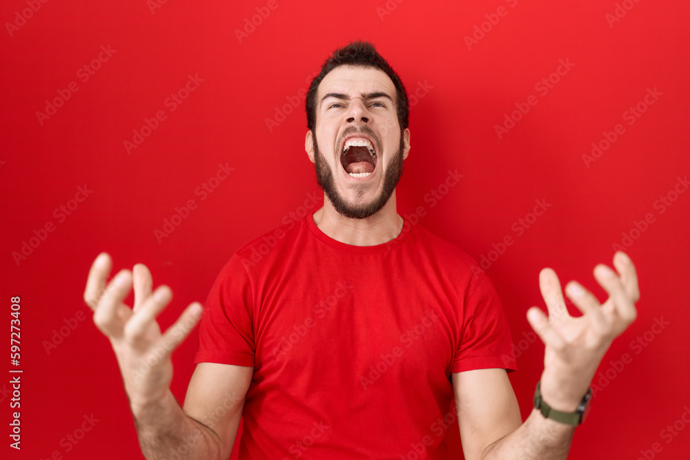 Young hispanic man wearing casual red t shirt crazy and mad shouting and yelling with aggressive expression and arms raised. frustration concept. Young hispanic man wearing casual red t shirt crazy and mad shouting and yelling with aggressive expression and arms raised. frustration concept.