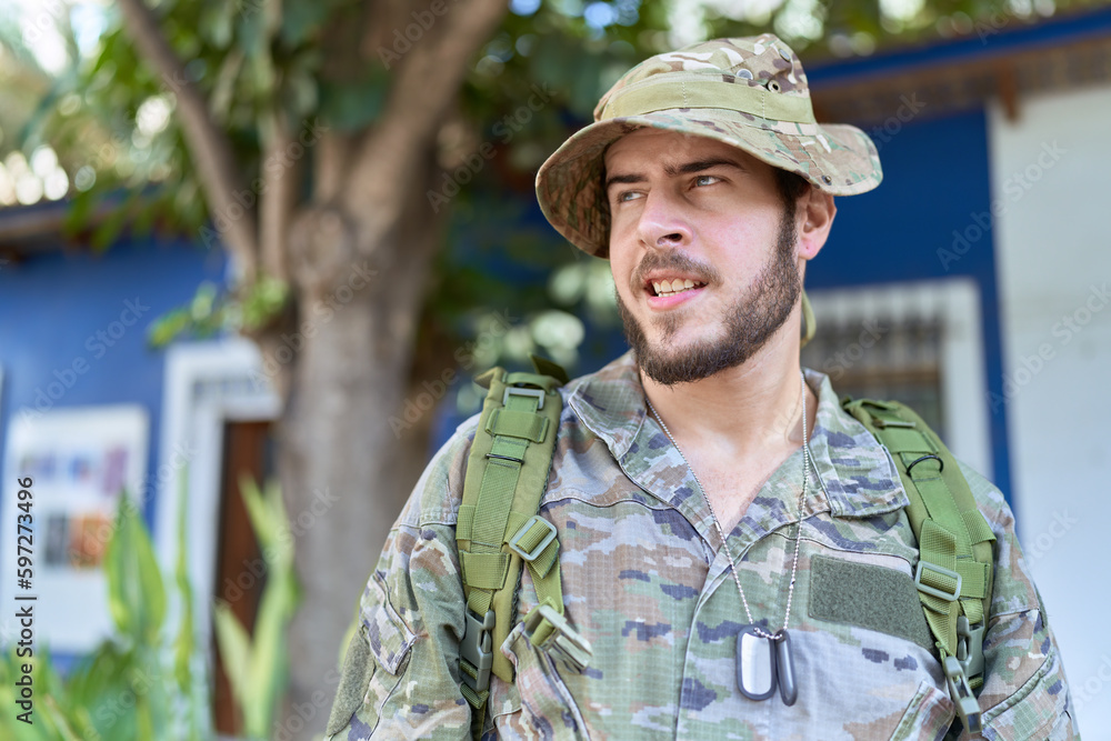Young hispanic man wearing camouflage army uniform outdoors looking away to side with smile on face, natural expression. laughing confident.