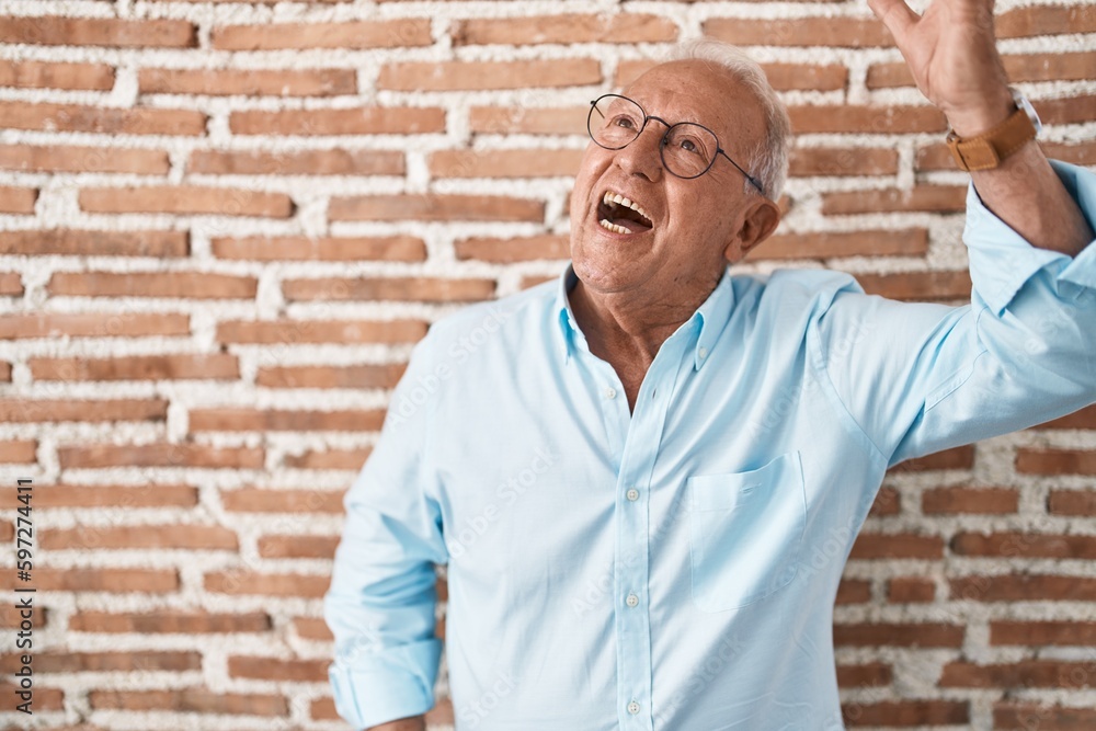 Senior man with grey hair standing over bricks wall dancing happy and cheerful, smiling moving casual and confident listening to music Senior man with grey hair standing over bricks wall dancing happy and cheerful, smiling moving casual and confident listening to music