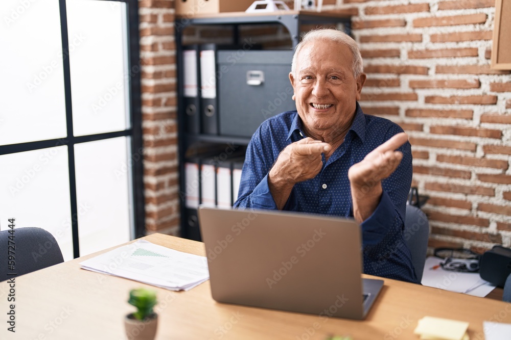 Senior man with grey hair working using computer laptop at the office amazed and smiling to the camera while presenting with hand and pointing with finger. Senior man with grey hair working using computer laptop at the office amazed and smiling to the camera while presenting with hand and pointing with finger.
