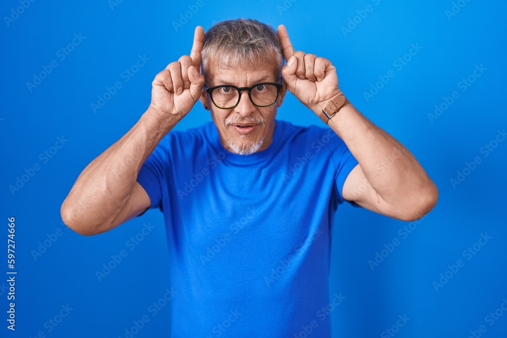 Hispanic man with grey hair standing over blue background doing funny gesture with finger over head as bull horns Hispanic man with grey hair standing over blue background doing funny gesture with finger over head as bull horns