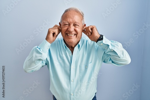 Senior man with grey hair standing over blue background covering ears with fingers with annoyed expression for the noise of loud music. deaf concept.