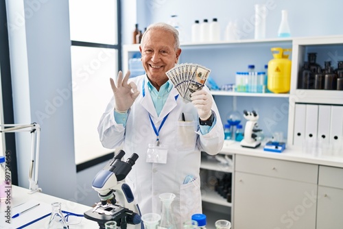Senior scientist with grey hair working at laboratory holding dollars doing ok sign with fingers, smiling friendly gesturing excellent symbol