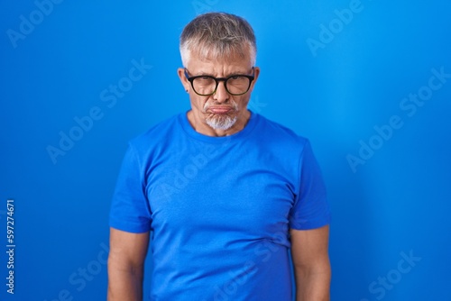 Hispanic man with grey hair standing over blue background skeptic and nervous, frowning upset because of problem. negative person.