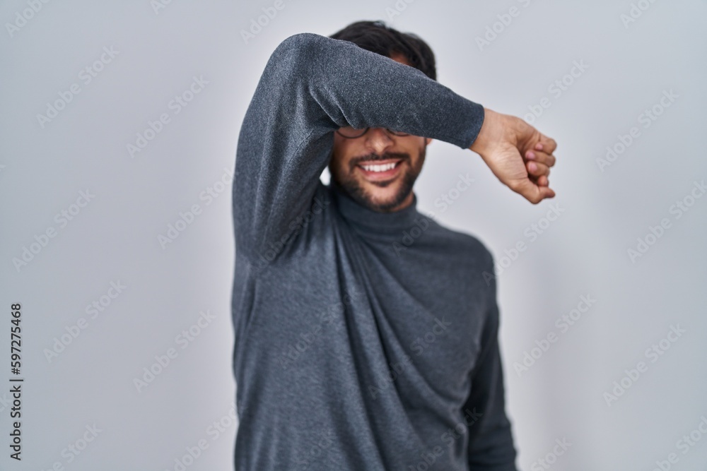 Handsome latin man standing over isolated background covering eyes with arm smiling cheerful and funny. blind concept. Handsome latin man standing over isolated background covering eyes with arm smiling cheerful and funny. blind concept.