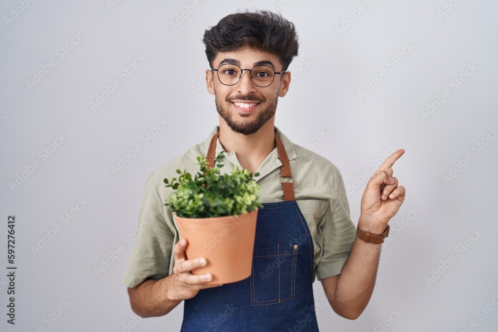 Arab man with beard holding green plant pot cheerful with a smile on face pointing with hand and finger up to the side with happy and natural expression Arab man with beard holding green plant pot cheerful with a smile on face pointing with hand and finger up to the side with happy and natural expression
