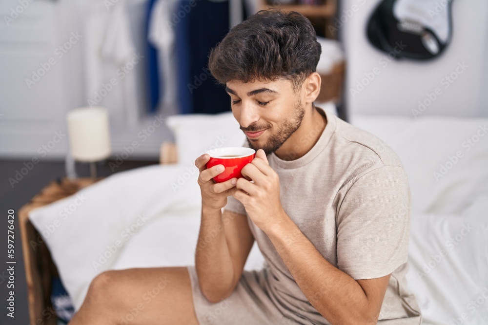 Young arab man drinking cup of coffee sitting on bed at bedroom