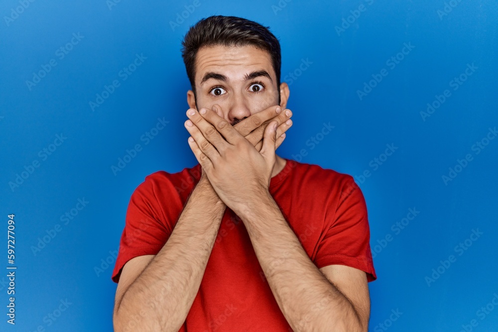 Young hispanic man with beard wearing red t shirt over blue background shocked covering mouth with hands for mistake. secret concept. Young hispanic man with beard wearing red t shirt over blue background shocked covering mouth with hands for mistake. secret concept.