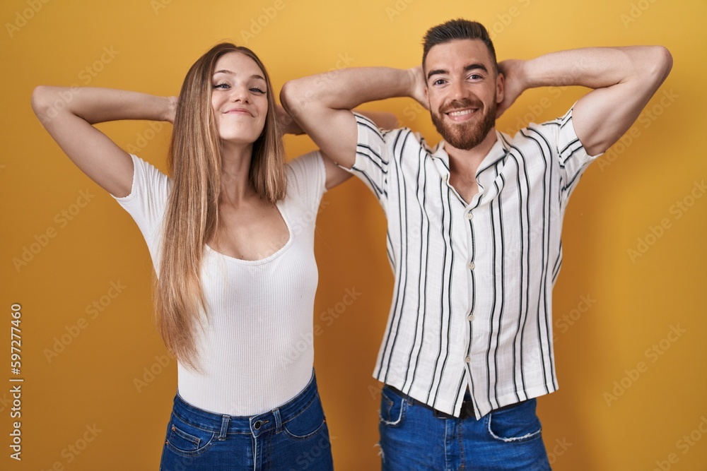 Young couple standing over yellow background relaxing and stretching, arms and hands behind head and neck smiling happy Young couple standing over yellow background relaxing and stretching, arms and hands behind head and neck smiling happy