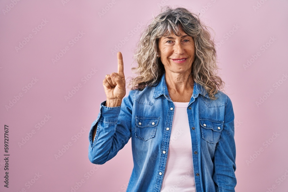 Middle age woman standing over pink background showing and pointing up with finger number one while smiling confident and happy. Middle age woman standing over pink background showing and pointing up with finger number one while smiling confident and happy.