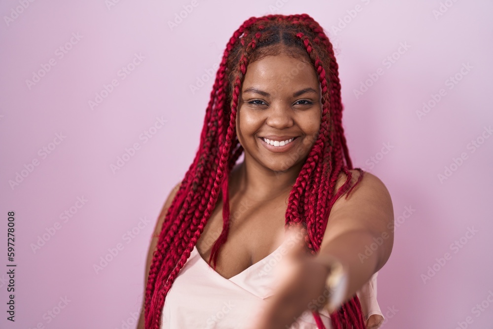 African american woman with braided hair standing over pink background smiling friendly offering handshake as greeting and welcoming. successful business. African american woman with braided hair standing over pink background smiling friendly offering handshake as greeting and welcoming. successful business.