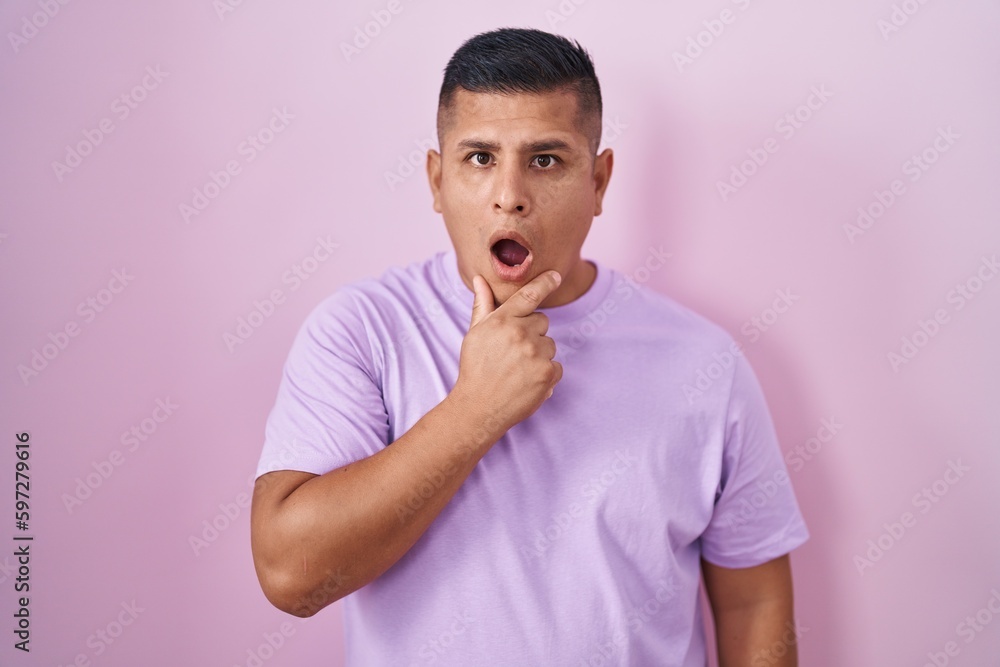 Young hispanic man standing over pink background looking fascinated with disbelief, surprise and amazed expression with hands on chin Young hispanic man standing over pink background looking fascinated with disbelief, surprise and amazed expression with hands on chin