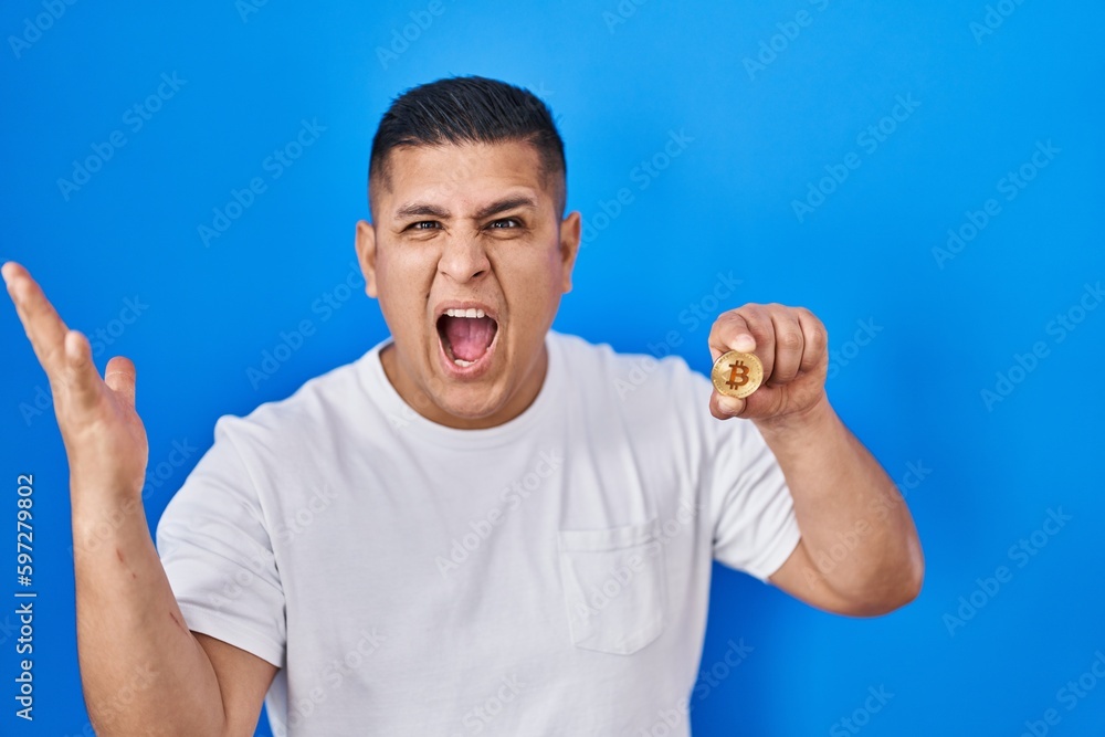 Hispanic young man holding virtual currency bitcoin celebrating victory with happy smile and winner expression with raised hands Hispanic young man holding virtual currency bitcoin celebrating victory with happy smile and winner expression with raised hands