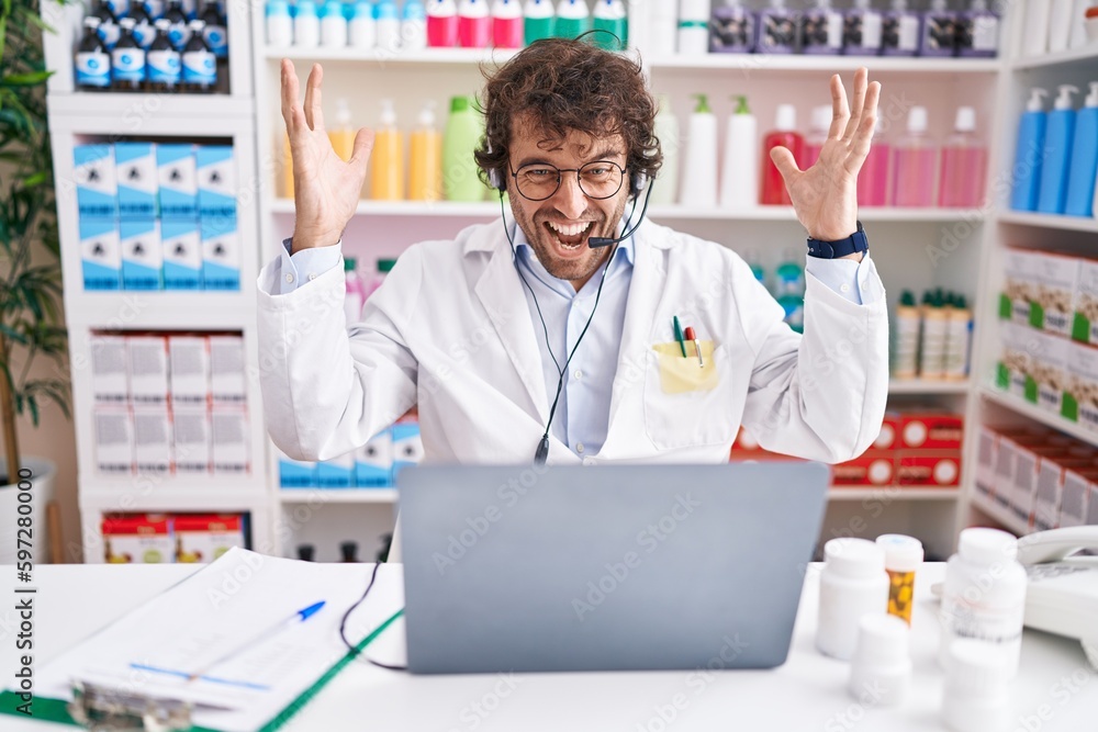 Hispanic young man working at pharmacy drugstore working with laptop celebrating victory with happy smile and winner expression with raised hands