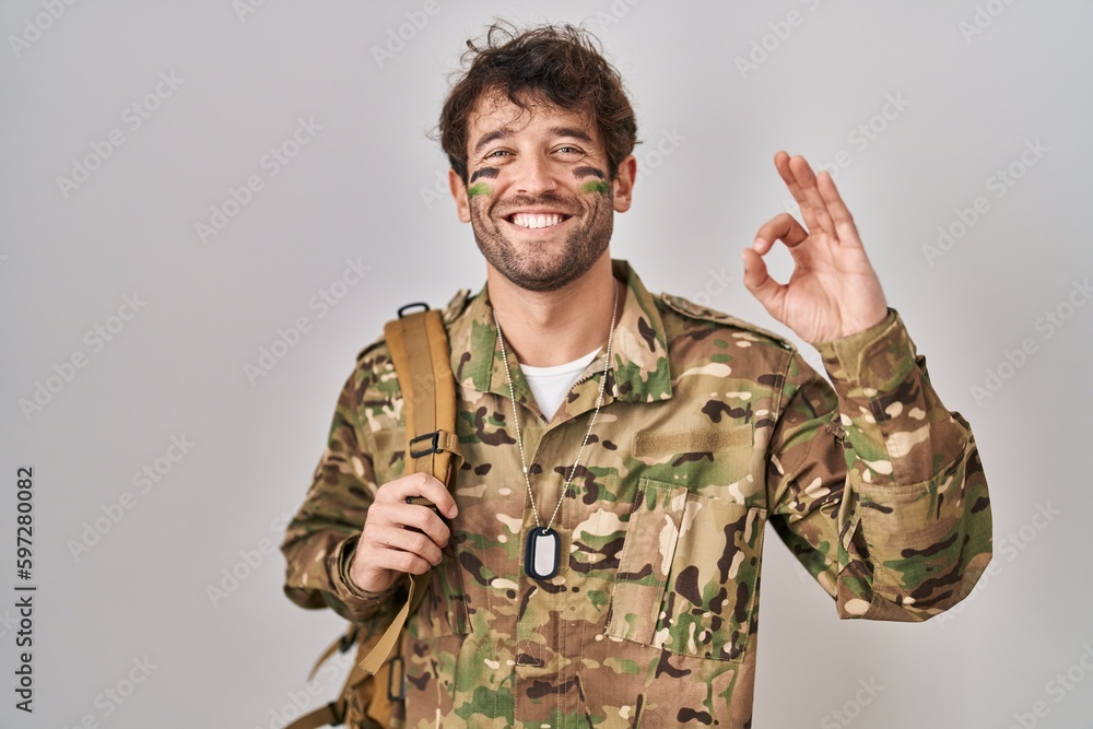 Hispanic young man wearing camouflage army uniform smiling positive doing ok sign with hand and fingers. successful expression. Hispanic young man wearing camouflage army uniform smiling positive doing ok sign with hand and fingers. successful expression.