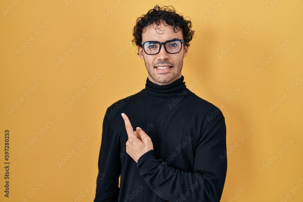 Fototapeta premium Hispanic man standing over yellow background pointing aside worried and nervous with forefinger, concerned and surprised expression