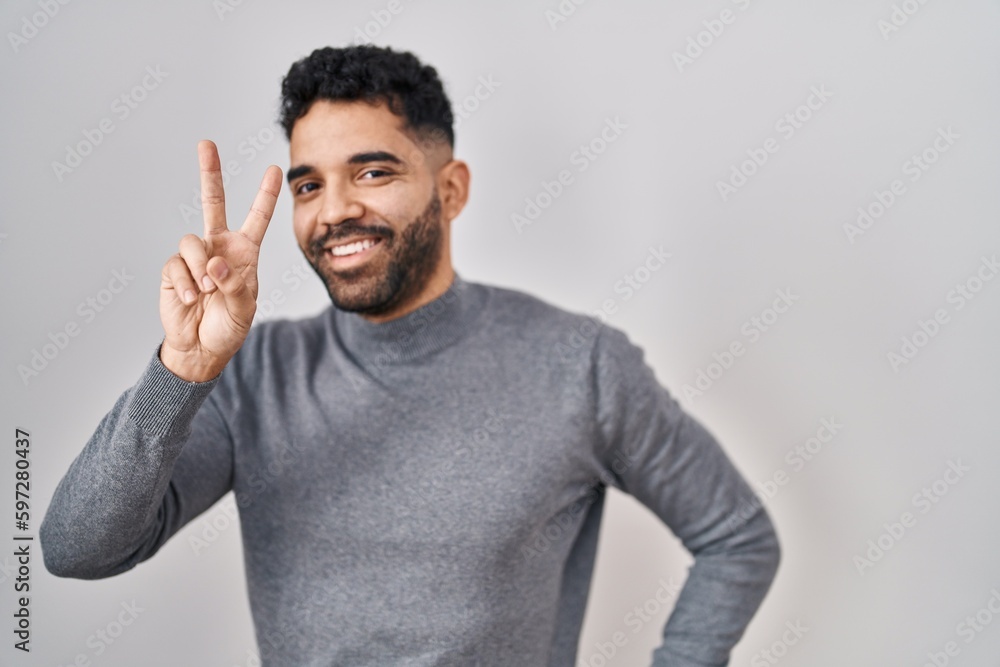 Hispanic man with beard standing over white background smiling looking to the camera showing fingers doing victory sign. number two. Hispanic man with beard standing over white background smiling looking to the camera showing fingers doing victory sign. number two.