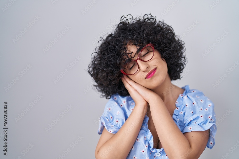 Young brunette woman with curly hair wearing glasses over isolated background sleeping tired dreaming and posing with hands together while smiling with closed eyes. Young brunette woman with curly hair wearing glasses over isolated background sleeping tired dreaming and posing with hands together while smiling with closed eyes.
