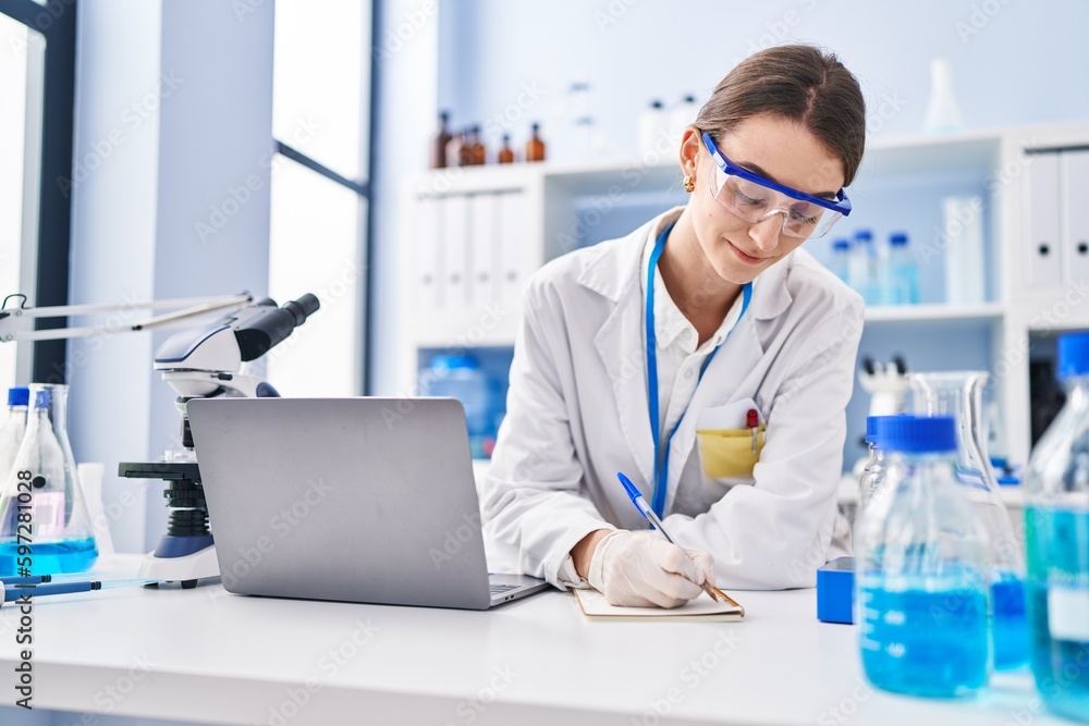 Young caucasian woman scientist using laptop writing on notebook at laboratory Young caucasian woman scientist using laptop writing on notebook at laboratory