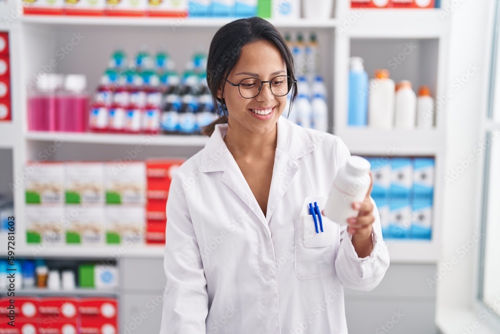 Young hispanic woman pharmacist smiling confident holding pills bottle at pharmacy