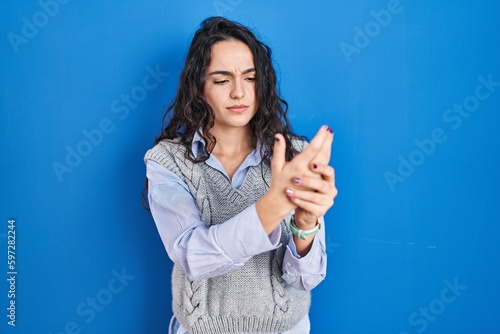 Young brunette woman standing over blue background suffering pain on hands and fingers, arthritis inflammation