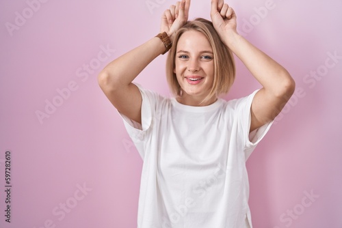 Young caucasian woman standing over pink background doing funny gesture with finger over head as bull horns