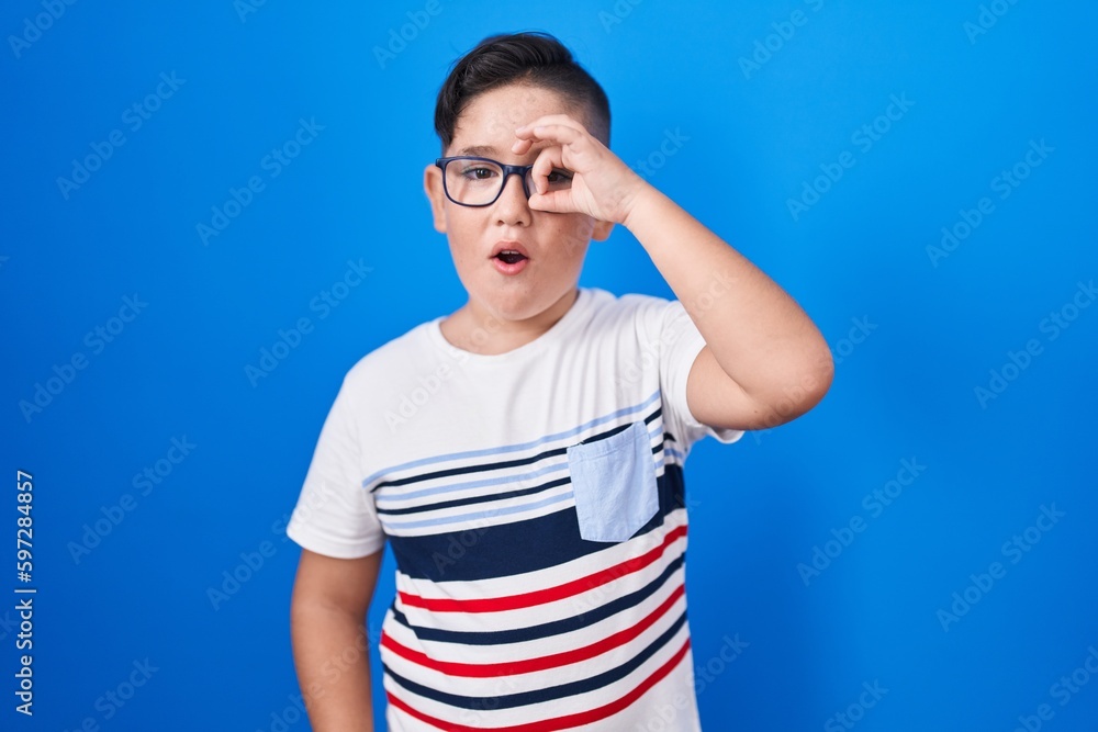Young hispanic kid standing over blue background doing ok gesture shocked with surprised face, eye looking through fingers. unbelieving expression. Young hispanic kid standing over blue background doing ok gesture shocked with surprised face, eye looking through fingers. unbelieving expression.