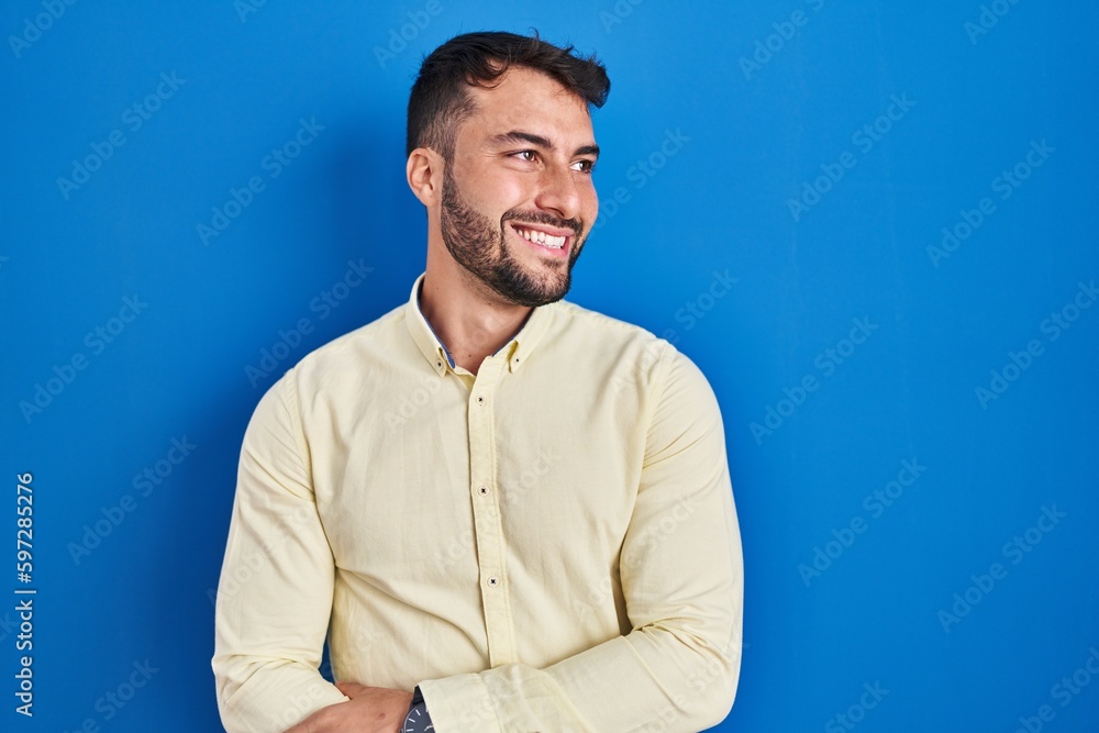 Obraz premium Handsome hispanic man standing over blue background looking away to side with smile on face, natural expression. laughing confident.