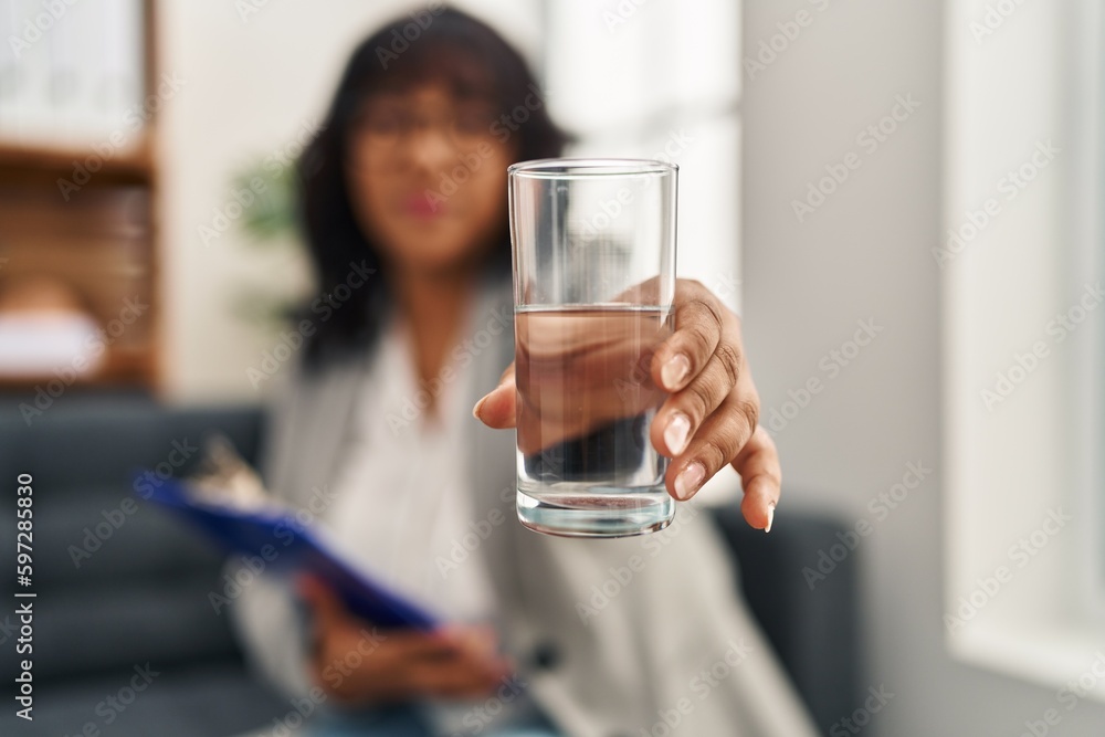Young beautiful latin woman psychologist smiling confident offering glass of water at psychology clinic