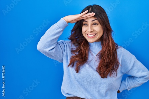 Hispanic young woman standing over blue background very happy and smiling looking far away with hand over head. searching concept.