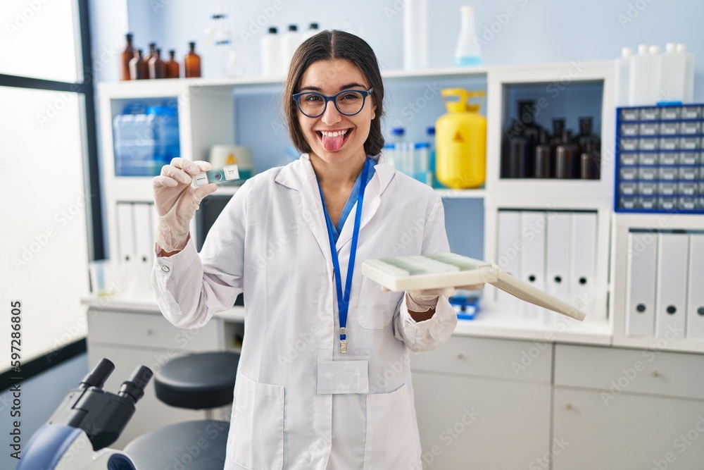 Young hispanic woman working at scientist laboratory with blood samples sticking tongue out happy with funny expression. Young hispanic woman working at scientist laboratory with blood samples sticking tongue out happy with funny expression.