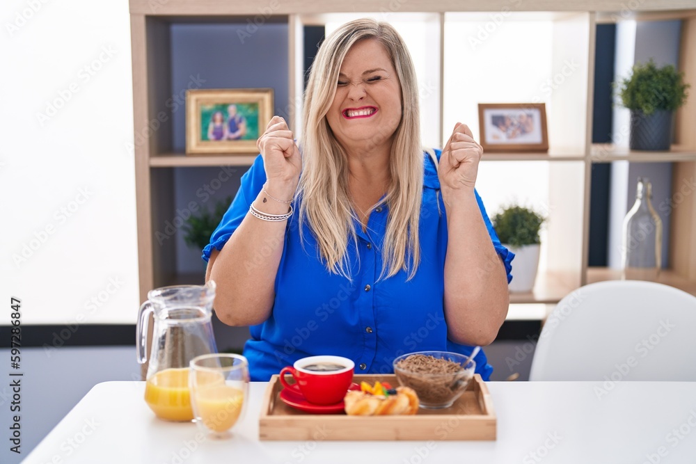 Caucasian plus size woman eating breakfast at home excited for success with arms raised and eyes closed celebrating victory smiling. winner concept. Caucasian plus size woman eating breakfast at home excited for success with arms raised and eyes closed celebrating victory smiling. winner concept.