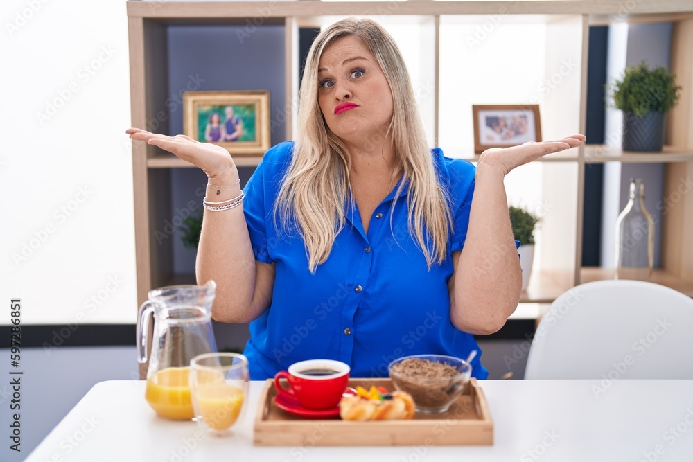 Caucasian plus size woman eating breakfast at home clueless and confused expression with arms and hands raised. doubt concept. Caucasian plus size woman eating breakfast at home clueless and confused expression with arms and hands raised. doubt concept.