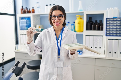 Young hispanic woman working at scientist laboratory with blood samples sticking tongue out happy with funny expression.