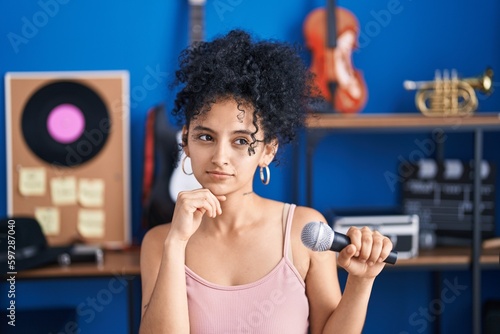 Hispanic woman with curly hair singing song using microphone at music studio serious face thinking about question with hand on chin, thoughtful about confusing idea