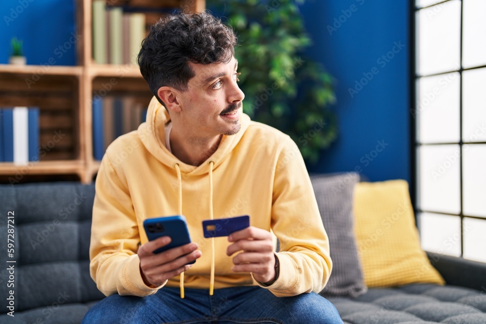 Young caucasian man using smartphone and credit card sitting on sofa at home Young caucasian man using smartphone and credit card sitting on sofa at home