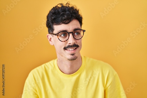 Hispanic man wearing glasses standing over yellow background looking away to side with smile on face, natural expression. laughing confident. © Krakenimages.com