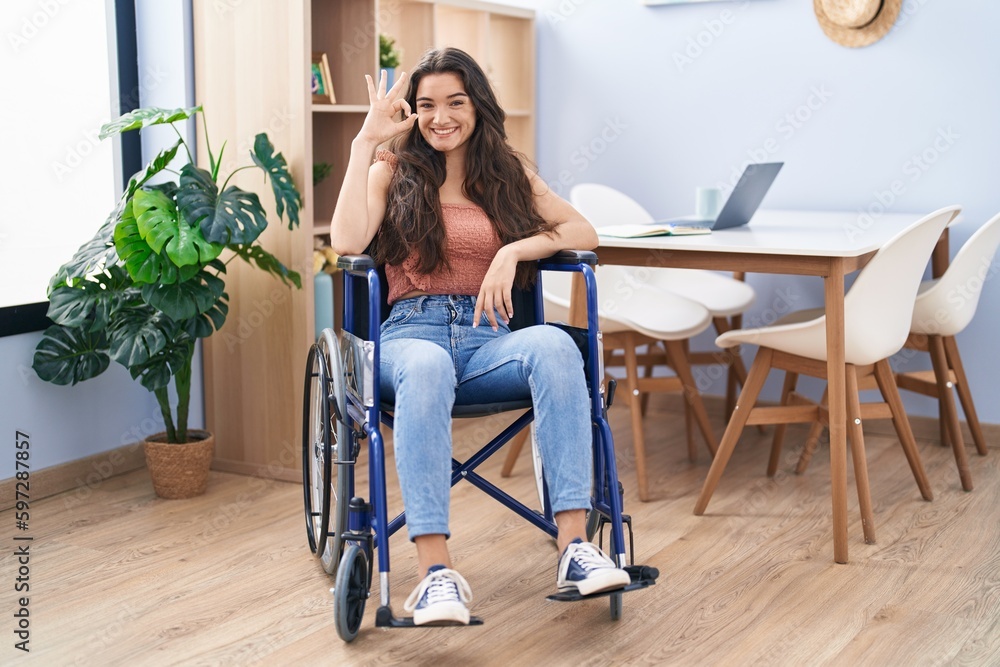 Young teenager girl sitting on wheelchair at the living room doing ok sign with fingers, smiling friendly gesturing excellent symbol Young teenager girl sitting on wheelchair at the living room doing ok sign with fingers, smiling friendly gesturing excellent symbol